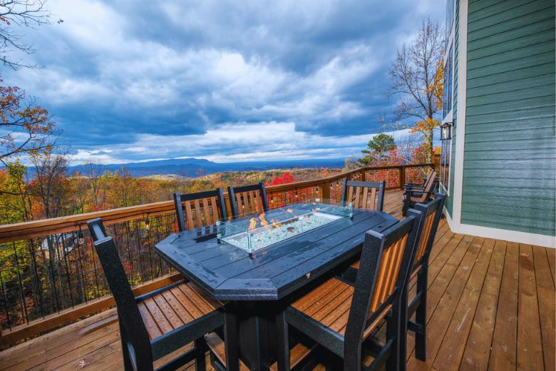 Wooden deck with a tall outdoor dining table featuring a built-in fire pit, surrounded by chairs and overlooking a colorful autumn forest and distant mountains under a dramatic cloudy sky.